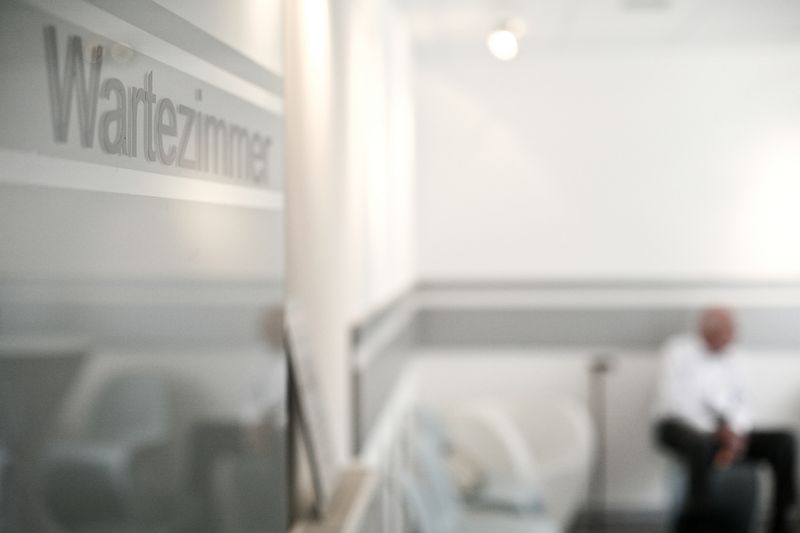 Glastür mit dem Schriftzug Wartezimmer, dahinter ein wartender Patient